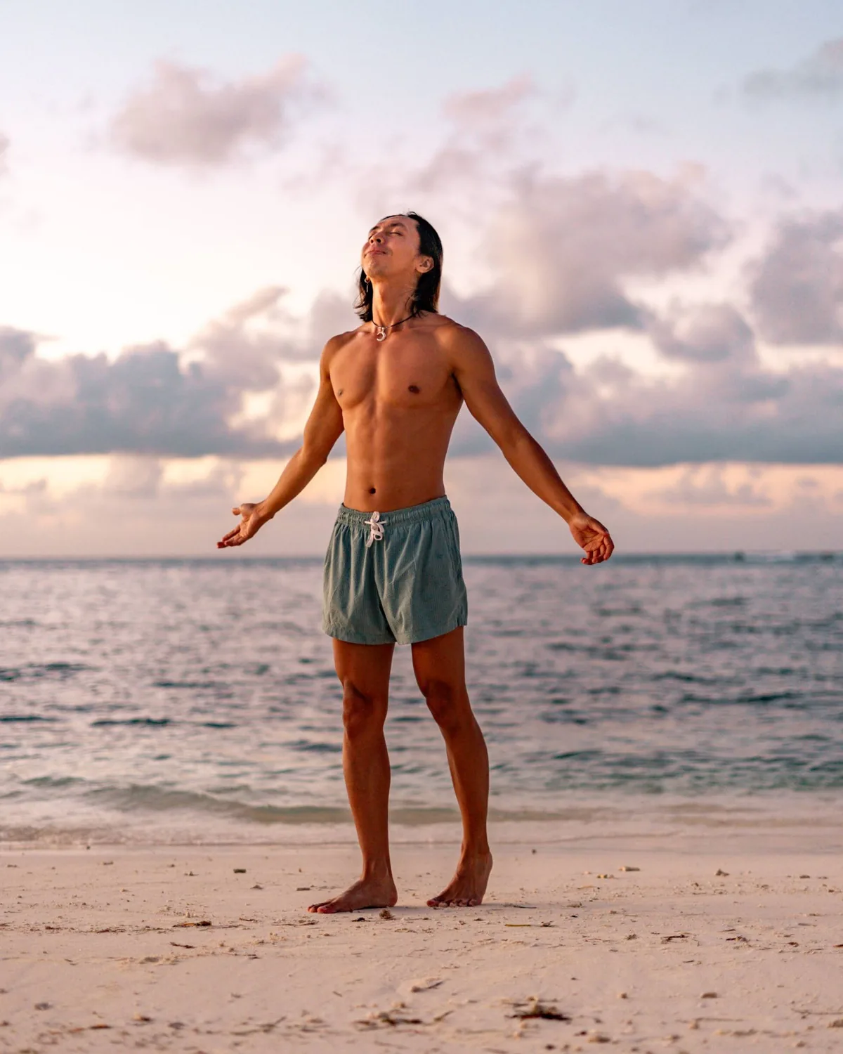 A man standing barefoot on the sand at sunrise, eyes closed and arms open to the sea