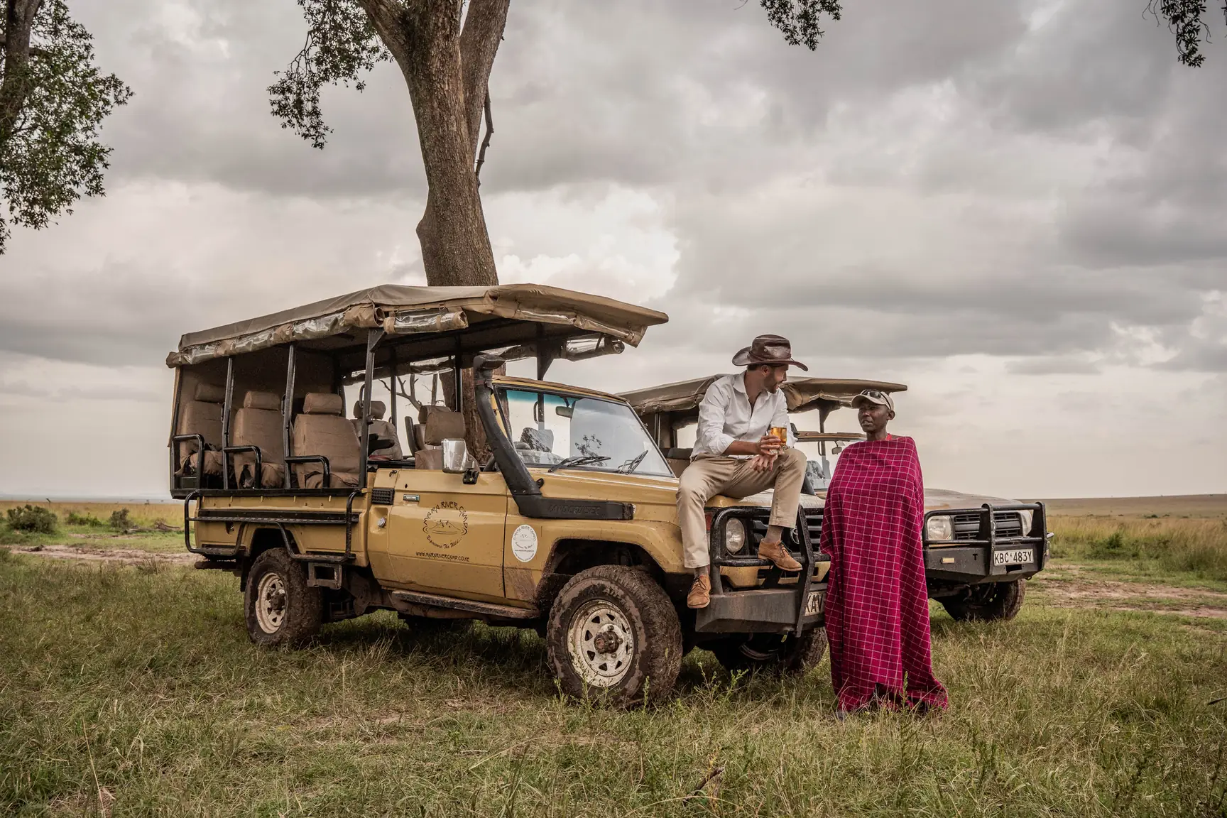 A Maasai guide in a red shuka standing beside a safari vehicle under a tree