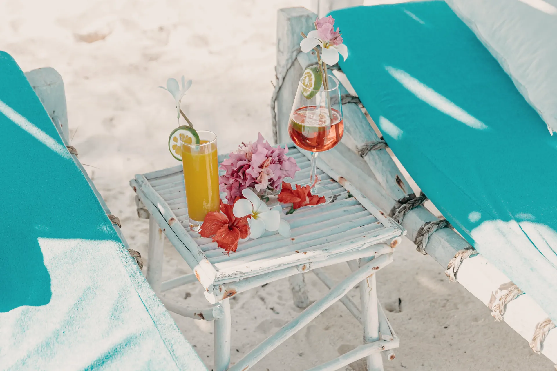 Two cold drinks with tropical flowers on a wooden table between sunbeds